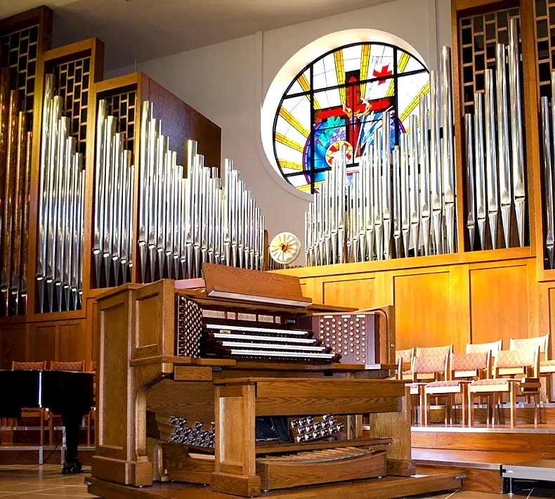 Allen organ console in a church sanctuary with a pipe facade and stained glass window
